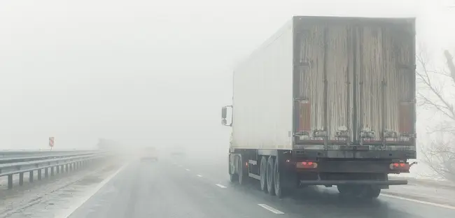 A semi-truck is driving in dangerous foggy conditions, highlighting driver negligence and reduced visibility hazards on the highway.
