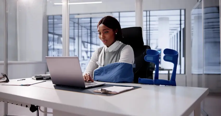 A woman with an arm injury in a sling is working at her desk and is filing a workers' compensation claim after a workplace accident in Georgia.