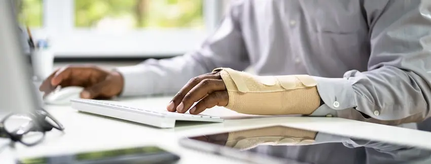 A man in a dress shirt with a wrist brace types on a keyboard with both hands at a desktop computer depicting an injured worker attempting to continue daily job duties while recovering and navigating the process of receiving workers' comp benefits in Georgia.