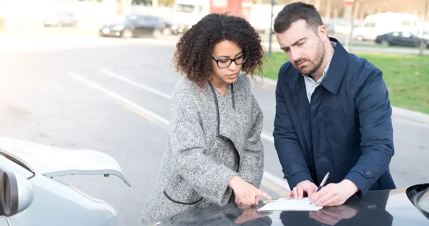 A woman wearing glasses and a man in a navy jacket review and sign paperwork on the hood of a damaged vehicle at the scene of a car accident documenting the incident before filing a claim with their health insurance company and with help from 1Georgia Personal Injury Lawyers.