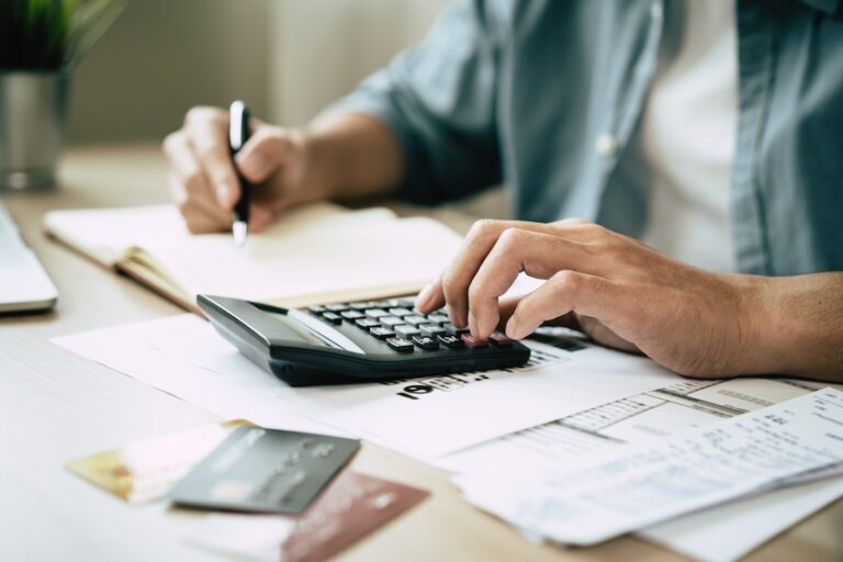 A person uses a calculator and pen to work through financial documents and bills spread across a desk alongside credit cards- calculating whether their coverage limits are enough to cover a car accident and all associated costs in Georgia.